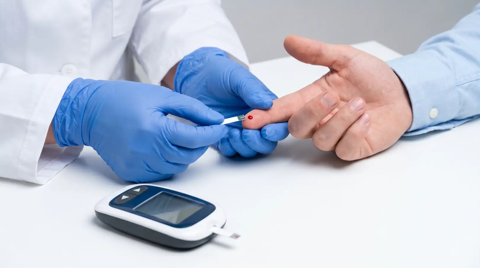 Gloved clinician collects a blood sample from a fingertip beside a glucose meter.