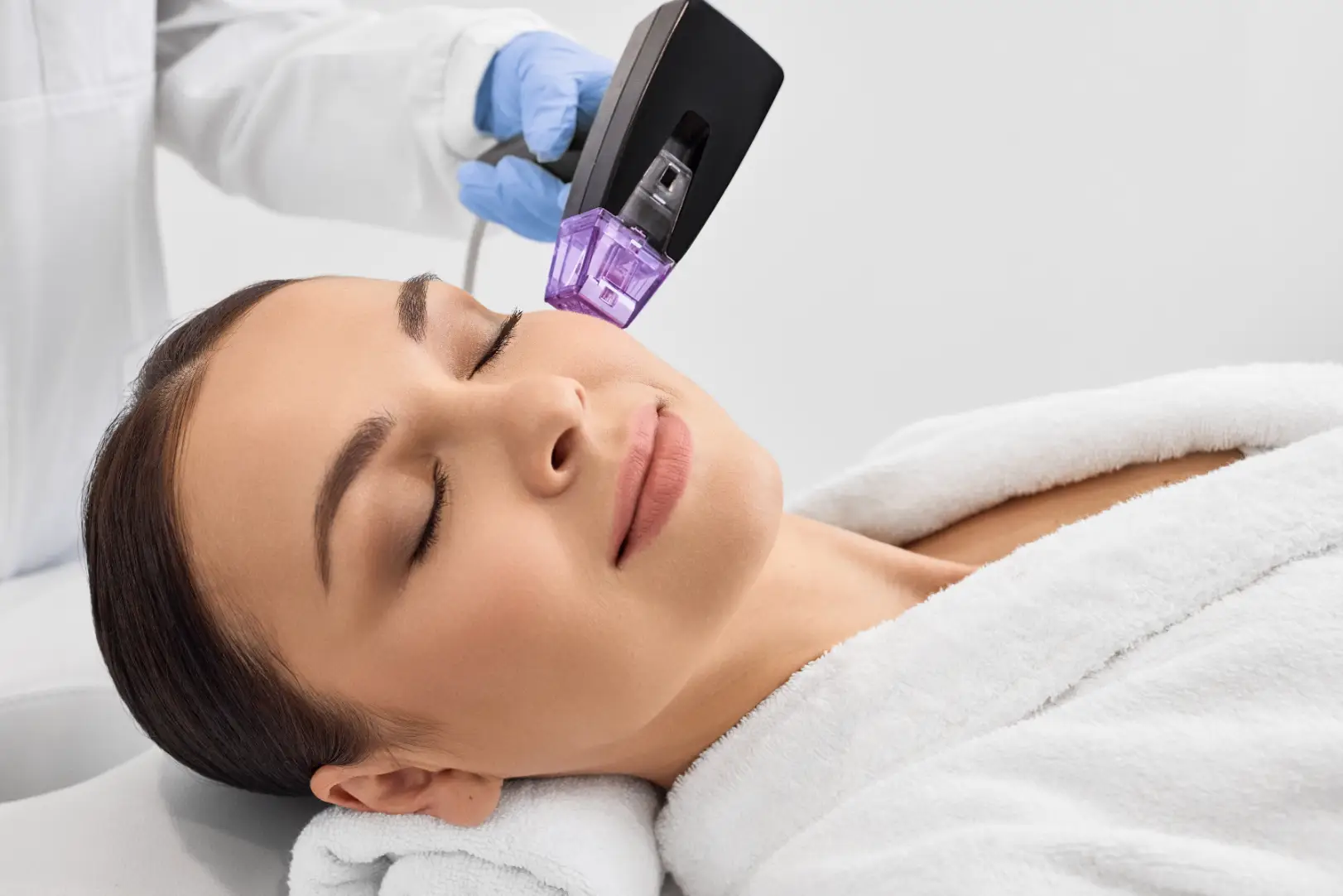 Woman lies with eyes closed while a gloved provider holds a handheld device against her cheek