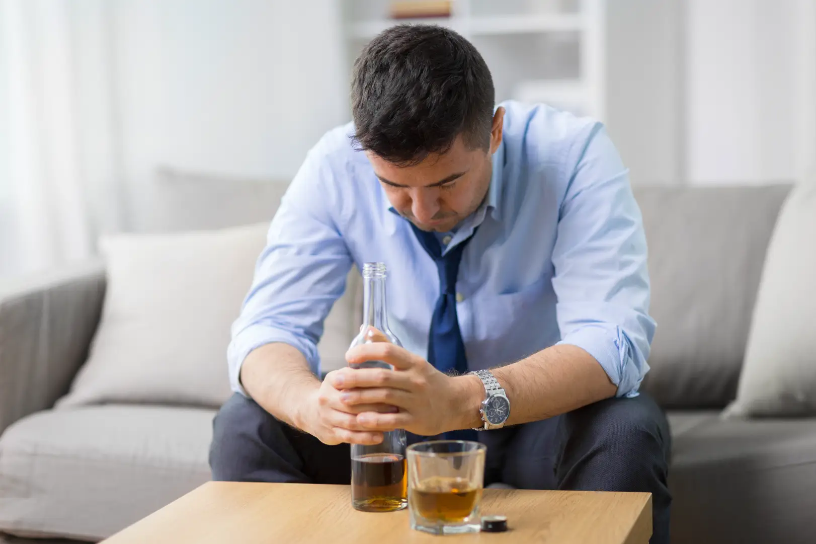 Man sits leaning forward on a sofa while holding a bottle, with a glass on the table.