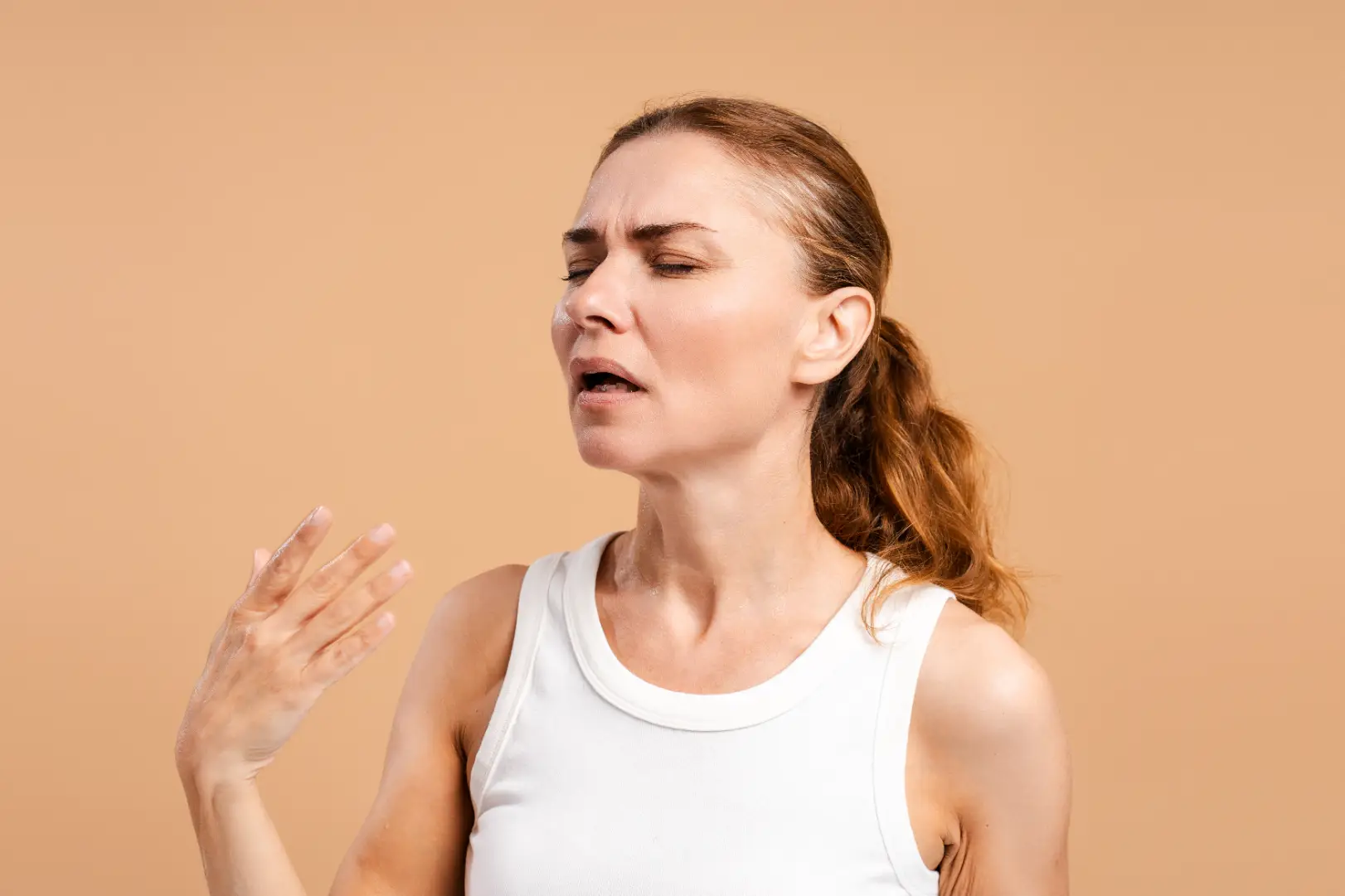 Woman in a white sleeveless top closes her eyes and raises one hand near her face against a peach background.