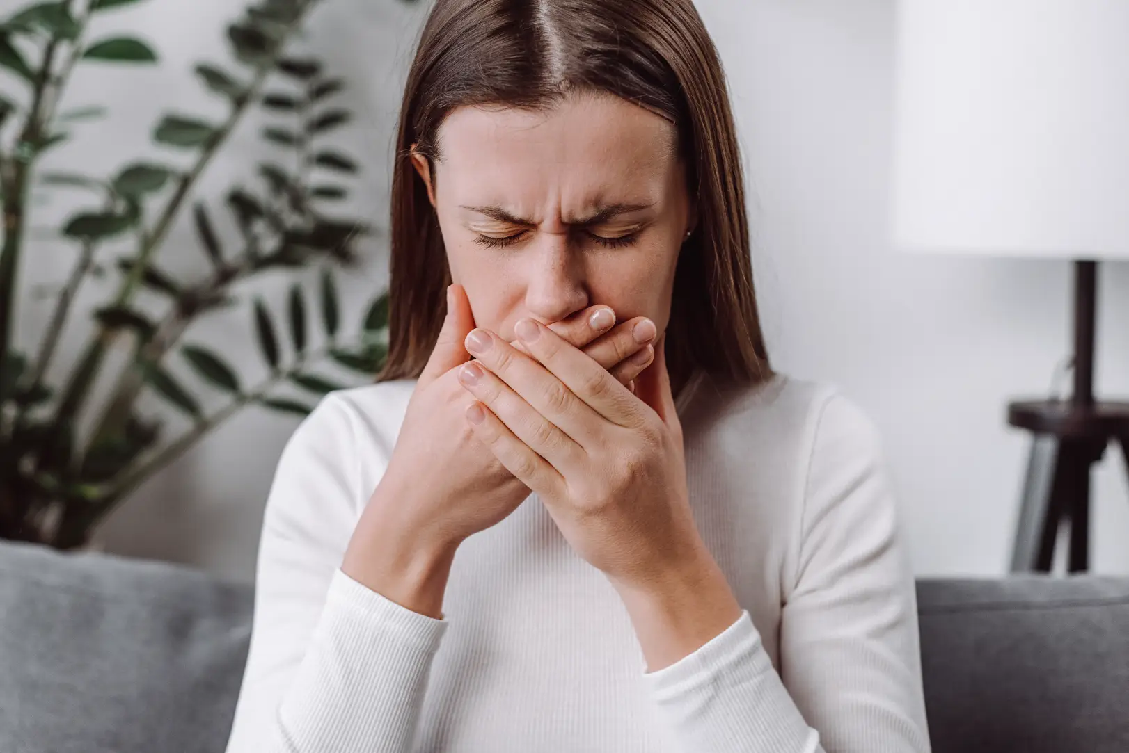 Woman sits indoors with her eyes closed and both hands covering her mouth.