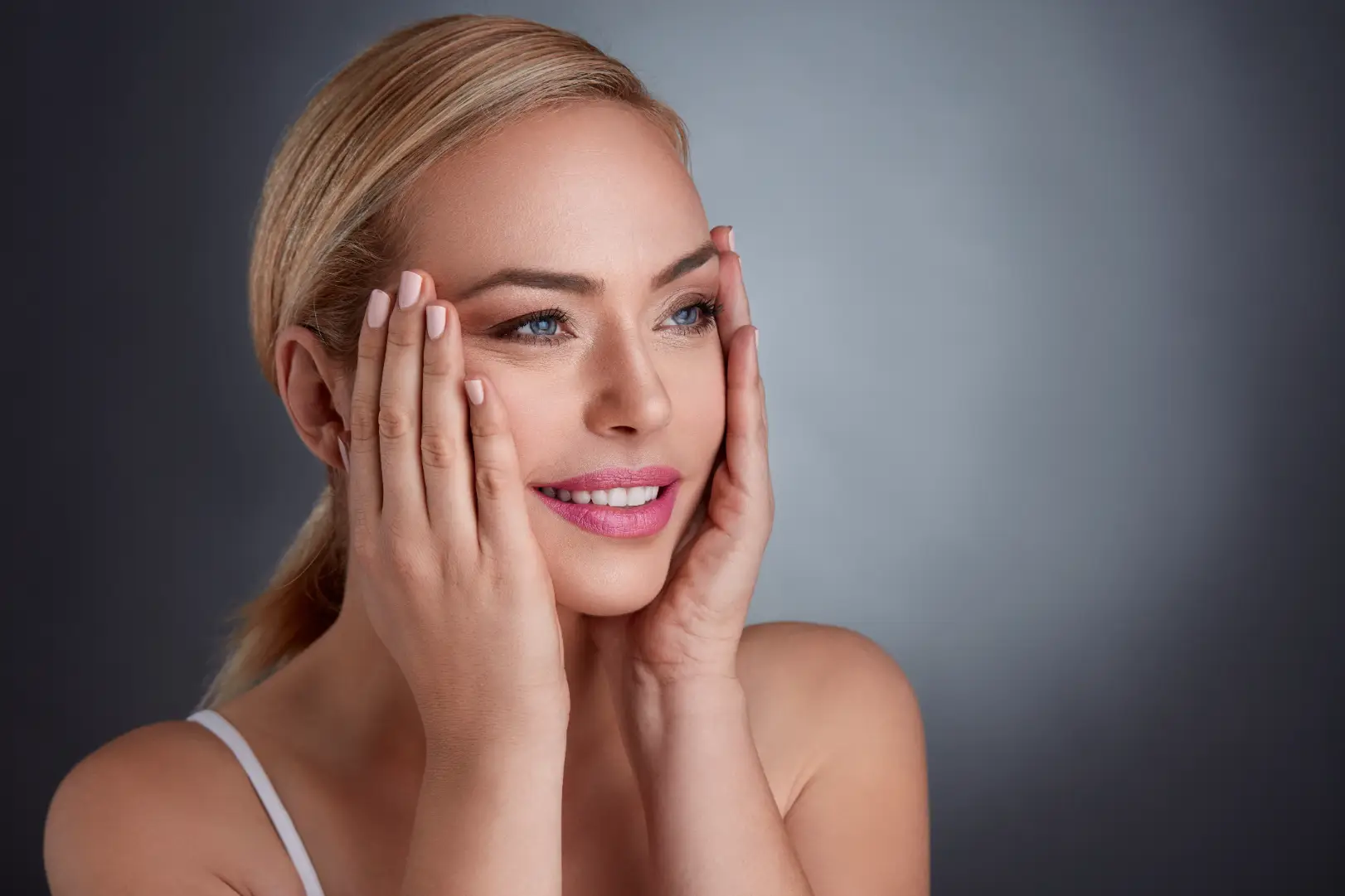 Smiling blonde woman gently touches both sides of her face against a dark gray studio background.